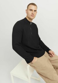 Schwarzer gerippter Pullover mit hohem Kragen und Detail mit halbem Reißverschluss, kombiniert mit hellbeigen Hosen. Das Model sitzt auf einem weißen Stuhl.