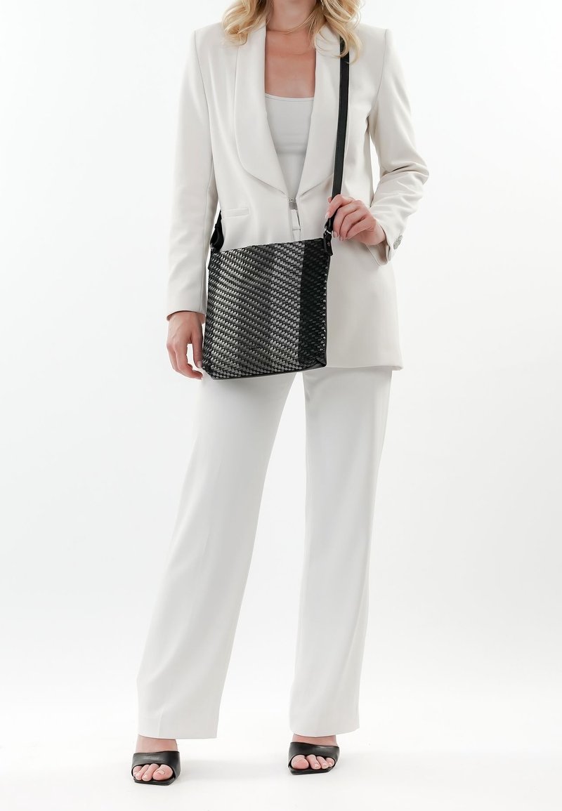 Femme portant un costume blanc et des talons ouverts noirs, tenant un sac bandoulière noir texturé avec une sangle d'épaule.
