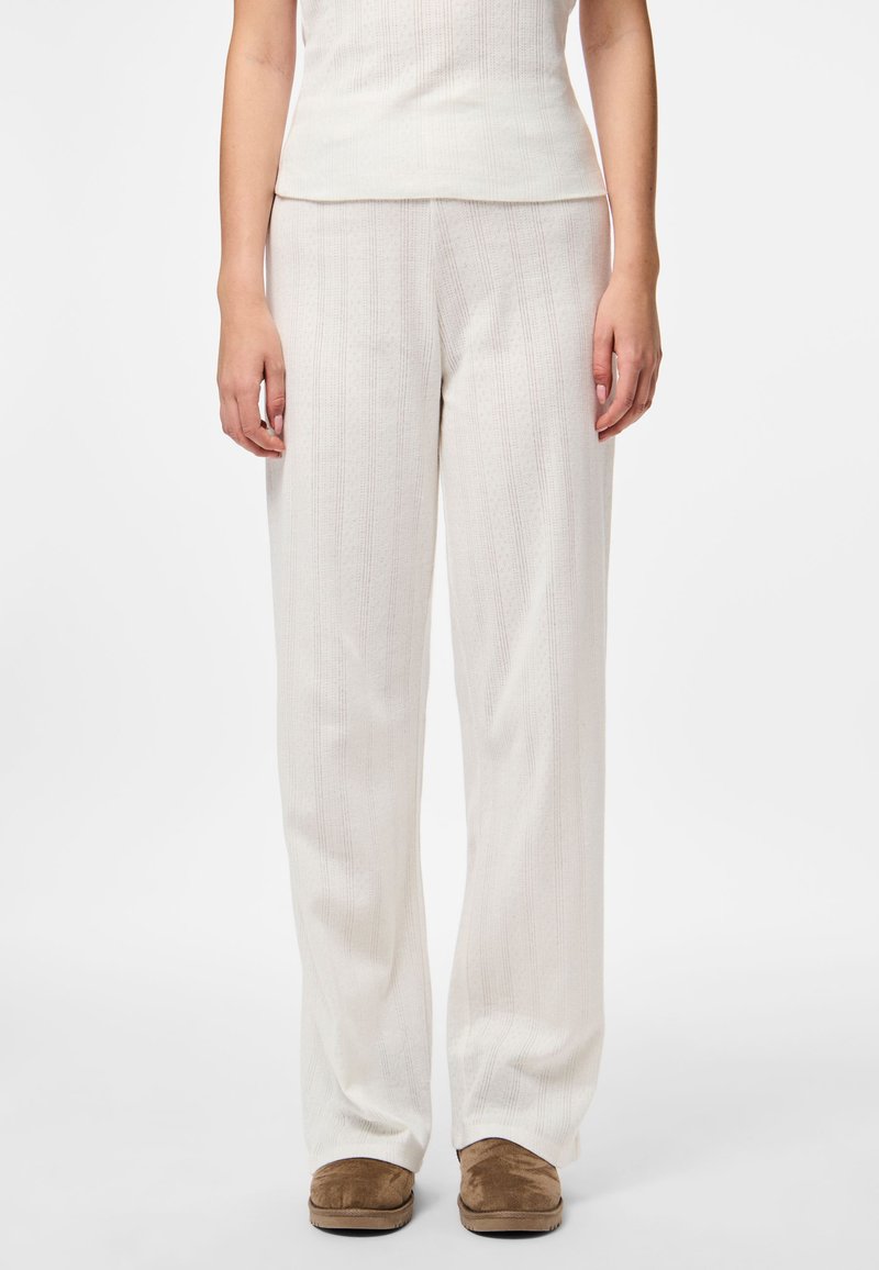 Lengvi, kreminės spalvos poilsio kelnės su vertikaliu dryžuotu tekstūru, plačiomis kojomis ir laisvu siluetu, derinamos su rudos spalvos šlepetėmis.