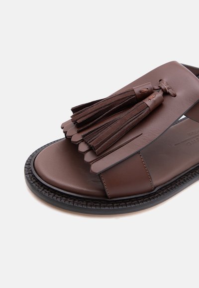 Sandale en cuir marron avec semelle noire, ornée de franges superposées et de deux pompons marron foncé sur la bride supérieure.