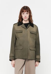 Casaco verde-azeitona com um colarinho de veludo preto, bolsos frontais e mangas longas. Textura suave com um design estruturado.