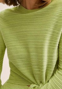 Długi sweter z rękawami w jaskrawym zielonym kolorze, o teksturowanym, żebrowanym wzorze. Posiada okrągły dekolt i detal w postaci węzła w talii.