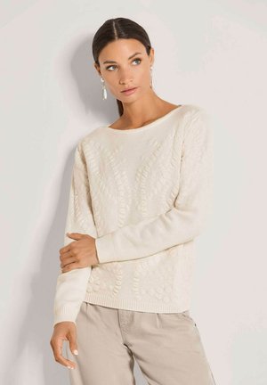 Vrouw met donker haar draagt een crème kabelgebreide trui en beige broek, kijkt opzij met één arm de andere bij de elleboog vasthoudend.
