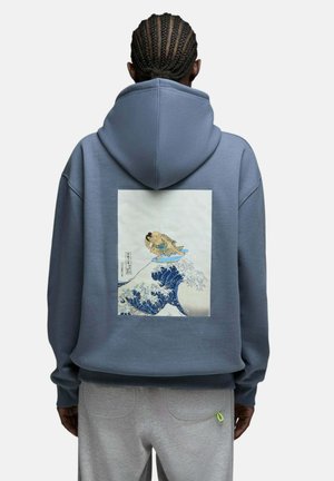 Personne portant un sweat à capuche bleu avec un graphisme de lutteurs de sumo surfant sur une grande vague océanique dans le dos, sur fond blanc.