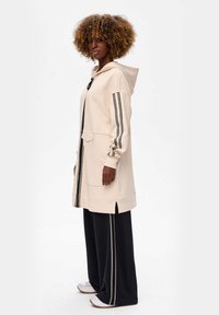 Vrouw met krullend haar, gekleed in een beigekleurige hooded zip-up jack met zwarte strepen en zwarte wijde broek met bijpassende strepen.