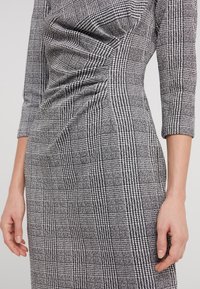 Robe cintrée à manches trois-quarts avec un motif pied-de-poule noir et blanc, présentant des fronces à l'avant et un tissu texturé.