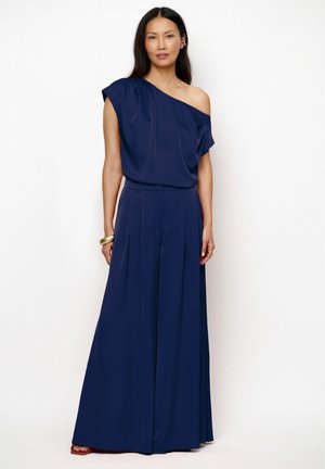 Femme portant un haut asymétrique bleu marine et un pantalon large assorti, debout devant un fond clair uni.