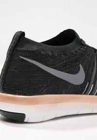 Scarpa sportiva nera in maglia con suola rosa oro, caratterizzata da un logo Nike grigio e superiore testurizzato. Design leggero con costruzione flessibile.