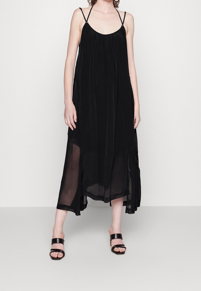 Femme portant une robe midi noire sans manches avec bretelles spaghetti et panneaux en tissu transparent, associée à des sandales à talons noires.