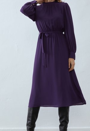 Kobieta ubrana w fioletową sukienkę midi z długim rękawem i paskiem w talii oraz czarne kozaki za kolano, stoi przy gładkiej białej ścianie.