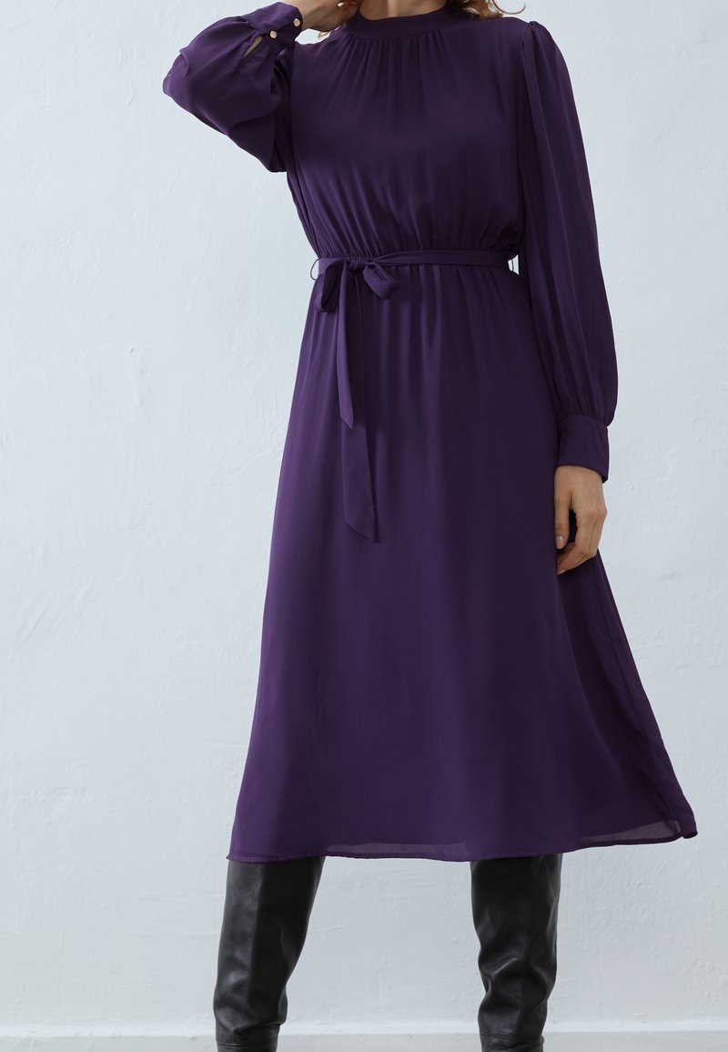 Femme portant une robe midi violette à manches longues avec une ceinture à la taille et des bottes noires montantes, debout contre un mur blanc uni.
