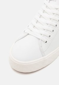 Valkoinen lenkkari lähikuvassa, jossa näkyy teksturoitu pohja, pyöreä kärki, valkoiset nauhat, metalliset reiät ja sileä synteettinen päällinen.