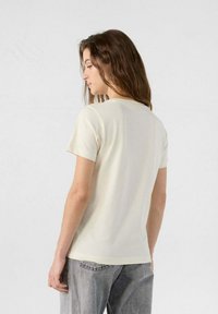 Camiseta de tirantes cortos de color crema claro con cuello redondo clásico, textura de tela suave y un diseño sencillo y limpio.