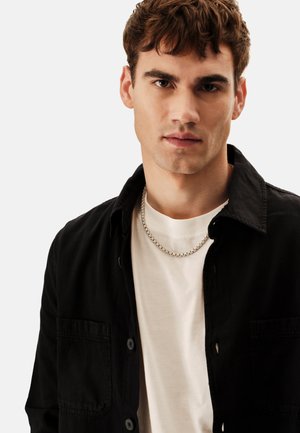 Zwarte overhemd met knopen en lange mouwen over een wit T-shirt; zilveren ketting; gladde stof; casual ontwerp met een borstzakdetail.