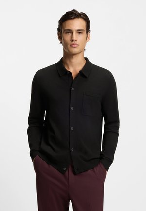 Homme portant une chemise noire boutonnée avec une poche poitrine et un pantalon rouge foncé, debout les mains dans les poches devant un fond uni.