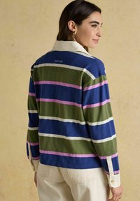 Sieviete, kas stāv šķērsām, ģērbusies garām svītrainām krekla ar zilām, zaļām, rozā un baltām svītrām un krēmkrāsas biksēs uz bēša fona.