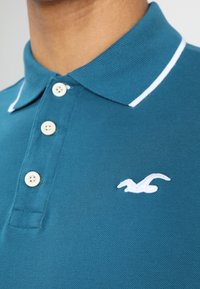 Turkoosi poolopaita, joka on valmistettu teksturoidusta kankaasta, valkoisilla raidallisilla kauluksilla, jossa on kolme nappia ja pieni brodeerattu logo.