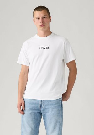 Ung mand iført en hvid Levi's t-shirt og lyseblå jeans står mod en ensfarvet lys baggrund.