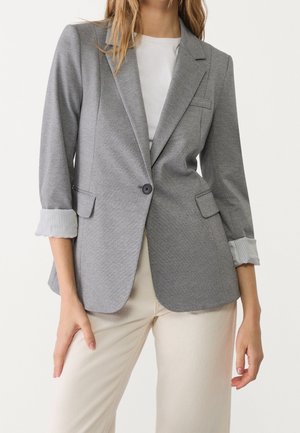 Vrouw die een lichtgrijze blazer met één knoop draagt, met opgerolde mouwen, een wit top en crèmekleurige broek tegen een effen achtergrond.