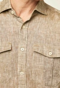 Chemise en lin marron clair avec un col, deux poches frontales et des fermetures à boutons. Tissu texturé avec un motif de tissage subtil.