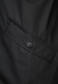 Musta vedenpitävä kangas, jossa on vaakasuora tasku ja pyöreä nappi, jossa lukee "RAIN". Sileä pinta ja minimalistinen muotoilu.