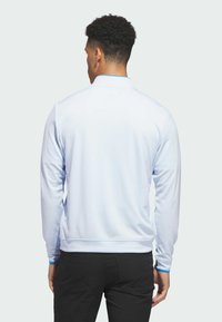 Jasnoniebieska koszulka polo z długim rękawem o teksturowanej tkaninie, z kołnierzykiem typu stójka i kontrastowym wykończeniem przy mankietach. Noszona z ciemnymi spodniami.