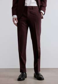Μπορντό παντελόνια ραμμένα από λείο ύφασμα, στενή εφαρμογή, με πτυχώσεις και τσέπες μπροστά. Συνδυασμένα με μαύρα loafers για αντίθεση.