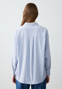 Camicia a righe blu e bianche con colletto, maniche lunghe e vestibilità ampia. Presenta una yoke posteriore e dettagli arricciati ai polsini.