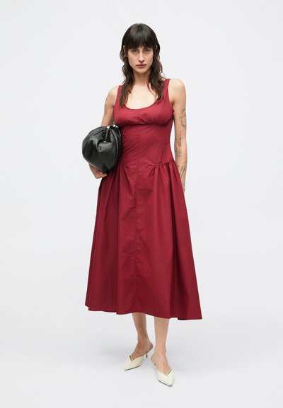 Κόκκινο βαμβακερό φόρεμα με εφαρμοστό μπούστο και φουσκωτή φούστα. Διαθέτει λαιμόκοψη σε σχήμα σέπια, λεπτές τιράντες και διακριτικές πιέτες στα πλάγια. Συνδυάζεται με μαύρη τσάντα.