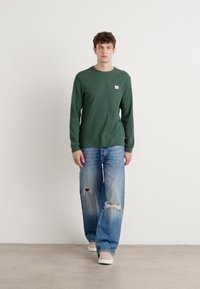 Donkergroen overhemd met lange mouwen en een gestructureerd patroon, gecombineerd met lichtblauwe versleten jeans en beige sneakers. Eenvoudig logo op het shirt.