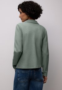Femme aux cheveux bouclés portant une veste texturée vert sauge et un jean bleu, debout contre un mur clair uni, de profil.