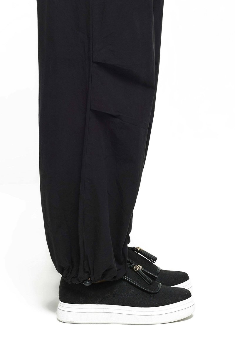 Parties basses des jambes portant un pantalon noir ample avec des poignets élastiques sur des chaussures noires à enfiler avec des semelles blanches et des détails à pompons.