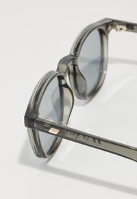 Ochelari de soare gri transparent cu lentile rectangulare, cadru curbat distinct și accente aurii pe braț. Text imprimerat pe templu.