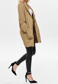 Beige oversized cardigan med forlommer, parret med sorte læderleggings og sorte højhælede sko, vist i profil.