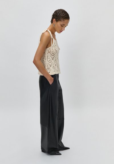 Γυναίκα με κοντά σγουρά μαλλιά, ντυμένη με αμάνικη πλεκτή μπλούζα εκρού και μαύρο δερμάτινο παντελόνι φαρδιάς γραμμής, στέκεται με τα χέρια στις τσέπες.