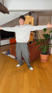 Model in a light gray sweater and loose gray trousers, standing with arms outstretched in a room with wood flooring and a potted plant.
