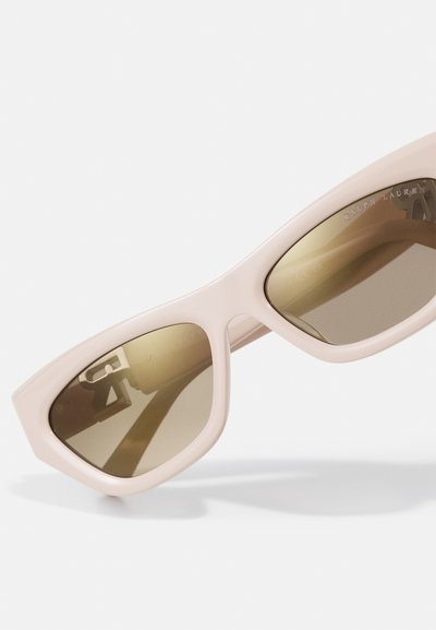 Sunglasses with a light pink plastic frame, angular cat-eye shape, and brown gradient lenses. Engraved logo detail visible on the lens.