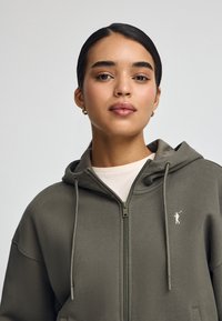 Felpa con zip verde oliva, dotata di un grande cappuccio, lacci e tasche frontali. Presenta un piccolo logo bianco sul lato sinistro del petto. Tessuto morbido.