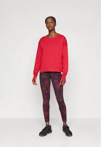 Sarkanā sporta džemperis ar garām piedurknēm, kopā nēsāts ar melnām pieguļošām legingām ar sarkaniem plankumiem. Melni sporta apavi papildina apģērbu.