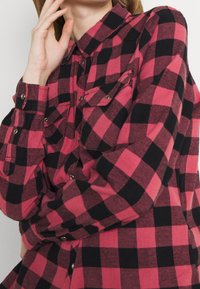 Red and black plaid flannel shirt with a button-down collar, two chest pockets, and silver snap buttons. Soft, textured fabric.