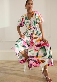 Robe à imprimé floral avec des manches bouffantes et une ceinture à nouer, présentant des motifs abstraits vibrants de rose, vert et orange. Portée avec des sandales à talons roses.