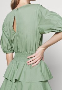 Nainen, jolla on yllään vaaleanvihreä mekko, jossa on puffihihat, kerätty vyötärö, peplum-helma ja avaus yksityiskohtana ylätakana. Käsi nojaa lantioon.