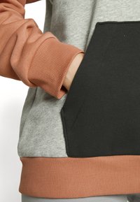 Šedá mikina z tkaniny s oranžovými rukávy, černou kapsou a žebrovaným lemem. Hladká textura, ležérní design.