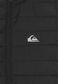 Giacca a vento nera in stile puffa con design trapuntato, chiusura frontale con zip e una vistosa toppa con logo argentato sul lato sinistro. Tessuto morbido.