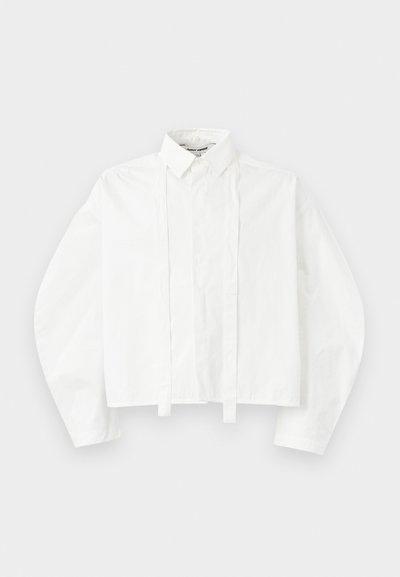 Camisa blanca de manga larga y cuello, con un diseño recortado. Presenta una tapeta frontal con lazos y un bordado de logo sutil en el pecho.
