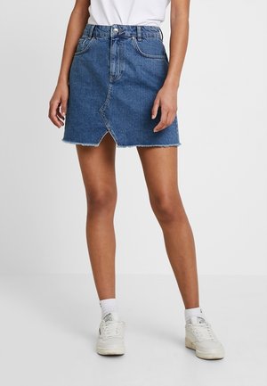 Mujer con una minifalda vaquera azul de dobladillo deshilachado y zapatillas blancas, de pie frente a un fondo blanco liso.