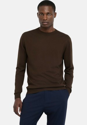 Maglione marrone a maniche lunghe realizzato in tessuto morbido e strutturato. Presenta una scollatura tonda e polsini a coste, abbinato a pantaloni blu navy.