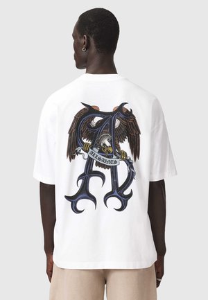 Homme de dos portant un T-shirt blanc avec un grand « A » stylisé et un graphisme d’aigle tenant une banderole indiquant « All Saints » dans le dos.