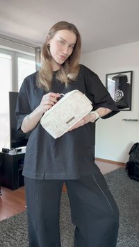 Jovem com cabelo comprido vestido de preto, segurando uma bolsa de cosméticos bege com padrão floral numa sala de estar.