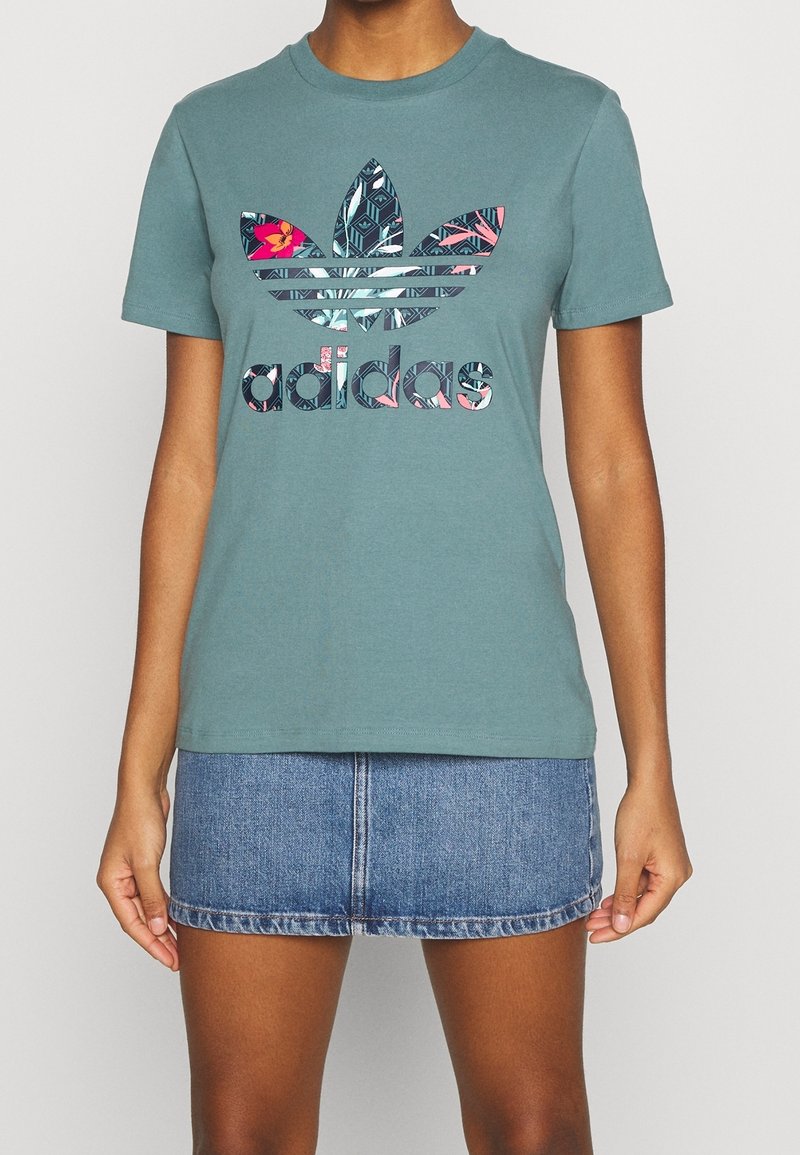 Teal bomullströja med en flerfärgad Adidas-logotyp med blommiga och geometriska mönster, i kombination med en jeanskjol.